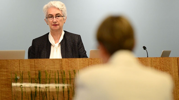 Justice Jennifer Coate sitting at a bench in the distance. The back of a second person is facing the Commissioner