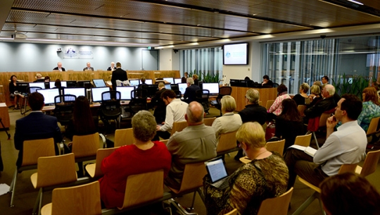 A hearing room with people facing forward sitting in chairs observing Commissioners sitting at a bench in the distance